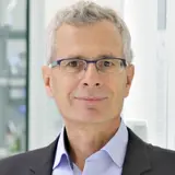 A middle-aged man with light gray hair and glasses is smiling at the camera. He is wearing a dark blazer over a light blue shirt and is standing in a well-lit environment, possibly an office or laboratory.