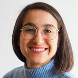 A smiling woman with shoulder-length hair wears round glasses and a blue turtleneck sweater. The background is light-colored, emphasizing her cheerful expression.