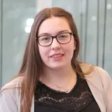 A young woman with long hair and glasses smiles while wearing a black shirt with lace details and a light cardigan. She is positioned in front of a background with large glass panels, suggesting a modern indoor environment.