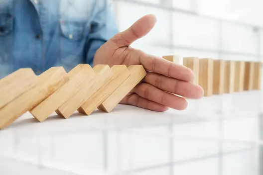 A hand is positioned in front of a series of tilted wooden blocks, resembling a domino setup. The image symbolizes the concept of interruption or prevention, possibly in a context related to research or innovation, reflecting themes of stability and control.