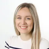 A woman with long, straight blonde hair is smiling at the camera. She is wearing a light-colored, striped shirt and has a welcoming expression. The background is plain and white, emphasizing her features.