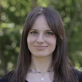 Ein junges Mädchen mit langen, dunklen Haaren lächelt in die Kamera. Sie trägt ein schwarzes T-Shirt und eine bunte Kette um den Hals. Der Hintergrund zeigt unscharf grüne Bäume, was eine natürliche Umgebung vermittelt.