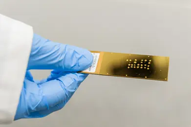 A gloved hand holds a shiny gold slide with a pattern of small dots. The slide features a label on one side. The background is neutral, emphasizing the scientific nature of the object.