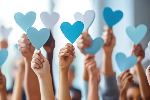 A diverse group of hands raises various shades of blue paper hearts in a show of support and unity. The hearts symbolize love and togetherness, creating a vibrant scene of collective enthusiasm and positivity.