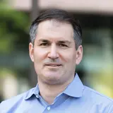 Porträt eines Mannes mit kurzen, dunklen Haaren, der ein hellblaues, kariertes Hemd trägt. Er steht vor einem unscharfen Hintergrund mit grünen Pflanzen. Der Mann lächelt leicht und blickt direkt in die Kamera.