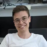 Ein junger Mann mit Brille lächelt freundlich. Er trägt ein weißes T-Shirt und sitzt in einem modernen Büro. Im Hintergrund sind Büroelemente wie Monitore und Pflanzen zu sehen.