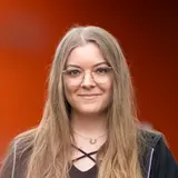 Eine lächelnde Frau mit langen, blonden Haaren steht an einem Geländer. Sie trägt eine schwarze Bluse und eine schwarze Jacke. Der Hintergrund ist orange und vermittelt eine freundliche Atmosphäre.