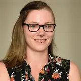 A smiling person with short hair and glasses poses for the photo. They wear a floral-patterned shirt. The background is light and neutral, creating a professional and friendly atmosphere.