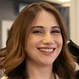 A young woman with medium-length brown hair and a warm smile is looking at the camera. She is wearing a white top and a delicate necklace. The background is slightly blurred, suggesting an indoor setting.