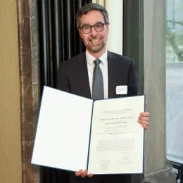Paul Martini Prize for therapeutic vaccinations against brain tumors A man in a suit is smiling while holding an official certificate. He stands indoors, with a dark backdrop. The certificate, mostly blank, indicates a formal recognition or award, possibly related to his professional achievements.