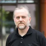 Porträt eines Mannes mit grauem, langen Haar und einem Bart. Er trägt ein schwarzes Poloshirt und steht vor einem unscharfen Hintergrund. Der Gesichtsausdruck ist neutral.