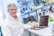 A scientist wearing a white lab coat and glasses is sitting at a lab bench and smiling at the camera. In front of him is a laptop displaying colorful microscopic images of tissue. In the background is a lab with shelves full of bottles, pipettes, and research equipment.