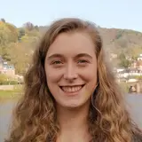 Eine junge Frau mit langen, lockigen Haaren lächelt freundlich in die Kamera. Sie trägt ein schwarzes Oberteil und einen blauen Blazer. Im Hintergrund sind Bäume und ein Fluss mit einer Brücke sichtbar, die eine malerische Szene ergänzen.