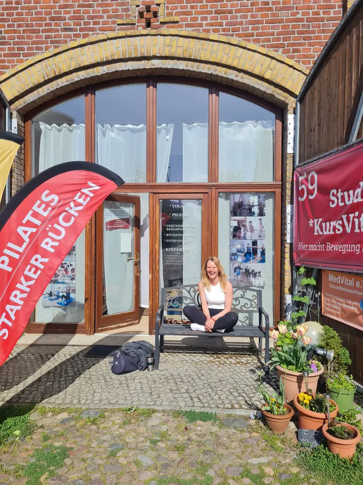 Stefanie Albrecht sitzt lächelnd auf einer Bank vor einem Eingang mit großen Fenstern