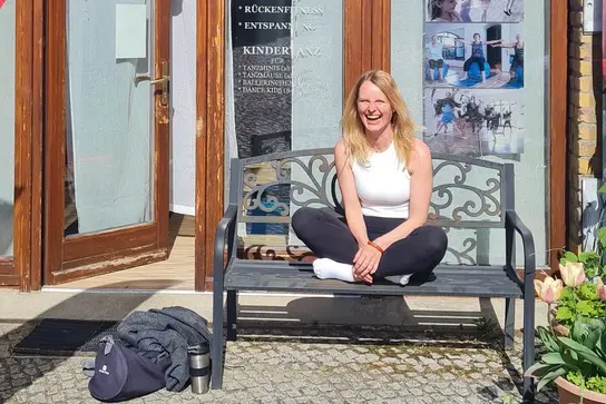 Stefanie Albrecht sitzt lächelnd auf einer Bank vor einem Eingang mit großen Fenstern