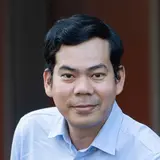 Ein Mann mit schwarzem, kurzem Haar steht an einem Geländer. Er trägt ein hellblaues Hemd und dunkelblaue Hose. Der Hintergrund zeigt verschwommene Gebäude und Grünflächen, die auf eine urbane Umgebung hinweisen. Der Mann hat einen freundlichen Ausdruck.