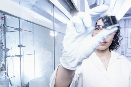 A scientist in a lab coat and white gloves holds up a small container filled with a dark substance. Behind her, laboratory equipment is visible, including glassware and scientific instruments. The setting is bright and modern, emphasizing a focus on research and experimentation.