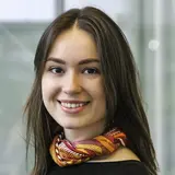A young woman with long, brown hair is smiling at the camera. She is wearing a black top and a colorful patterned scarf around her neck. The background appears light and unfocused, emphasizing her cheerful expression.
