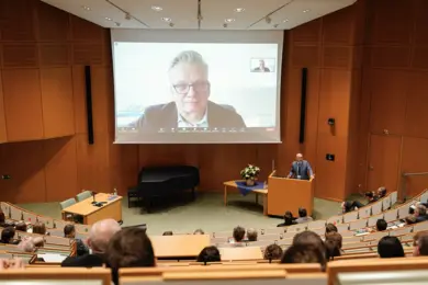 A speaker stands on stage in a conference hall, addressing an audience seated in rows. On a large screen behind them, a remote participant is engaged in a virtual presentation. The setting reflects a modern event focused on radiation therapy advancements in Heidelberg.