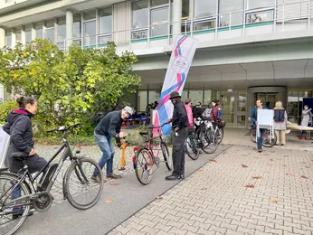 Employees wait in line with their bicycles outside a building. A man assists a colleague in inflating a bike tire.