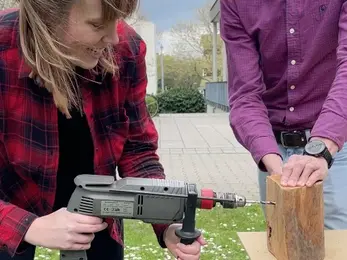 A staff member drills into a wooden log while a colleague holds it steady on a picnic bench outdoors.