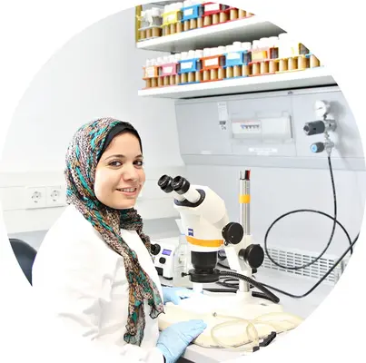 Sara, wearing a lab coat and gloves, sits next to a microscope.