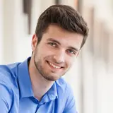 Ein junger Mann mit dunklen Haaren und einem gepflegten Bart trägt ein hellblaues Hemd. Er lächelt freundlich in die Kamera. Der Hintergrund ist unscharf und zeigt einen hellen Innenraum.