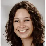 A young woman with curly brown hair smiles brightly at the camera. She wears a light-colored tank top, and her expression conveys warmth and friendliness. The background is a neutral gray, focusing attention on her joyful demeanor.