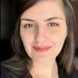 Porträt einer jungen Frau mit langen, dunklen Haaren und einem freundlichen Lächeln. Sie trägt ein schwarz gestreiftes Oberteil und eine dezente Halskette. Der Hintergrund ist unscharf, wodurch ihr Gesicht im Fokus steht.