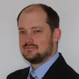 Ein Mann mit kurzem, braunem Bart und braunen Haaren blickt in die Kamera. Er trägt ein dunkles Sakko und ein hellblaues Hemd mit einer gestreiften Krawatte. Der Hintergrund ist neutral und hell.