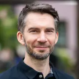 Ein lächelnder Mann mit kurzem, grauen Haar und einem Bart trägt ein schwarzes Polo-Shirt. Der Hintergrund zeigt unscharfe grüne und graue Elemente, die auf eine natürliche Umgebung hinweisen. Der Mann wirkt freundlich und entspannt.