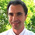 A smiling male doctor or healthcare professional wearing a white coat and a tie, standing outdoors in front of greenery. The image conveys a sense of professionalism and approachability.