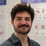 Ein Mann mit dunklen Haaren und Bart sitzt lächelnd vor wissenschaftlichen Postern in einem modernen Raum. Er trägt ein blaues Hemd und wirkt freundlich. Die Poster im Hintergrund enthalten Grafiken und Texte zu wissenschaftlichen Themen.