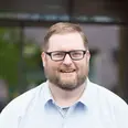 Ein lächelnder Mann mit Brille und einem karierten Hemd steht im Freien. Der Hintergrund zeigt unscharfe Bäume und Gebäude. Sein Gesichtsausdruck wirkt freundlich und einladend.