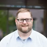 Ein lächelnder Mann mit Brille und einem karierten Hemd steht im Freien. Der Hintergrund zeigt unscharfe Bäume und Gebäude. Sein Gesichtsausdruck wirkt freundlich und einladend.
