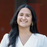 Eine lächelnde Frau steht an einem Geländer. Sie trägt eine weiße Bluse und einen farbigen Rock mit grafischem Muster. Im Hintergrund sind uneindeutige, unscharfe Farben zu sehen, die eine moderne Umgebung andeuten.