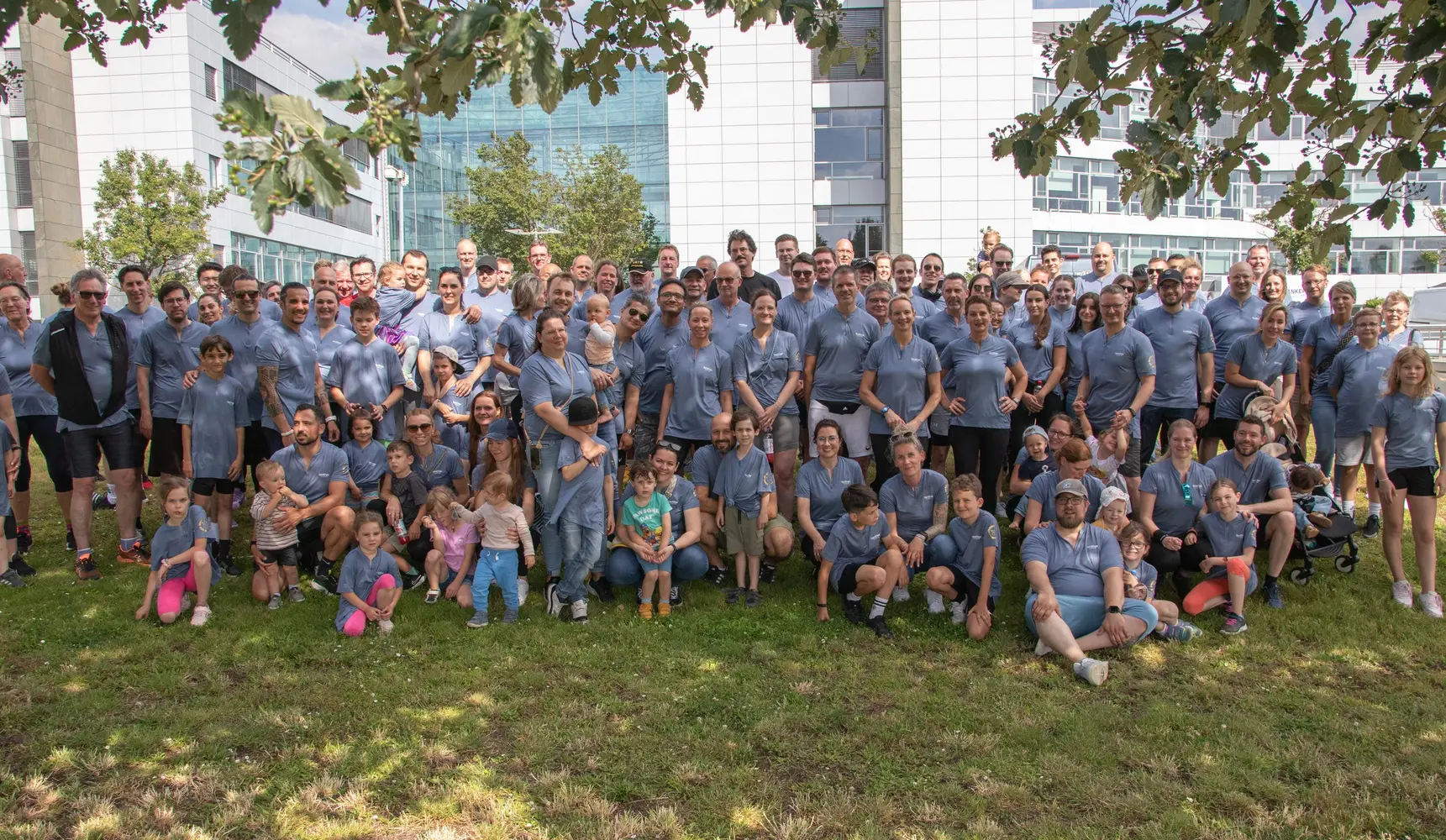 Eine große Gruppe von Menschen, darunter Erwachsene und Kinder, posiert gemeinsam im Freien vor einem großen Gebäude. 