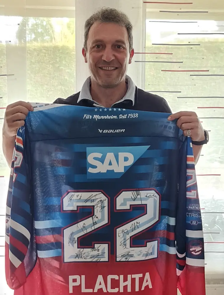 Ein Mann hält lächelnd ein Eishockey-Trikot mit der Nummer 22 in die Kamera. Das Trikot trägt den Namen „Plachta“ sowie das Logo von SAP. Auf dem Trikot sind mehrere Unterschriften der Spieler sichtbar.