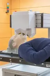 Research Topic Imaging and Radiooncology  A person lies on a medical imaging table, undergoing a scan. A machine positioned above their head appears to be scanning for diagnostic purposes. The environment is clinical, indicated by the smooth surfaces and neutral colors of the room.