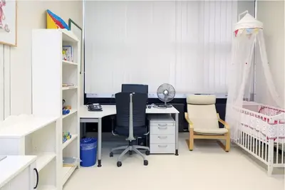 Das Bild zeigt ein Büro mit einem Schreibtisch, einem Computer, einem Ventilator und einem bequemen Sessel. Auf der rechten Seite befindet sich ein Kinderbett mit einem Moskitonetz, und links ist ein Regal mit Büchern und Spielzeug. Die Wand hat große Fenster mit Jalousien.
