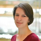 A young woman with short brown hair smiles warmly. She is wearing a deep red V-neck sweater and stands in a bright environment, suggesting a modern or academic setting. The background is softly blurred, highlighting her friendly expression.