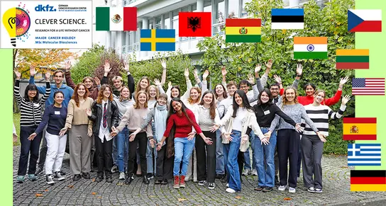 A diverse group of approximately 30 people stands outdoors in a cheerful pose, waving their arms. They are gathered in front of a building surrounded by greenery, showcasing a sense of collaboration and community.