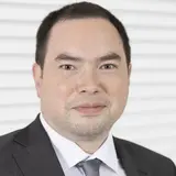 Ein Mann in einem dunkelgrauen Anzug steht vor einem weißen Hintergrund. Er hat kurze, dunkelbraune Haare und trägt eine hellgraue Krawatte. Sein Gesichtsausdruck ist freundlich und neutral.