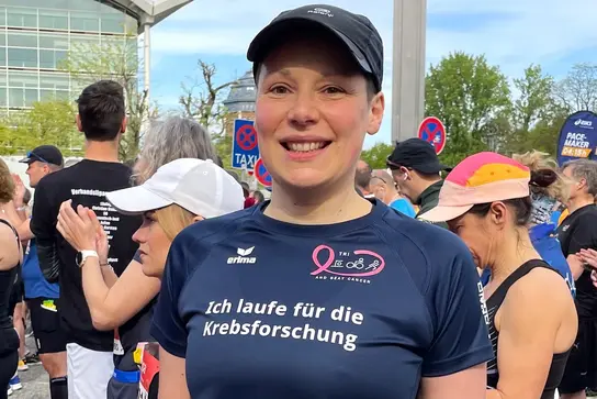 Michaela Thulke in Laufkleidung und mit Startnummer steht lächelnd inmitten von anderen Läufern. Auf ihrem T-Shirt steht: „Ich laufe für die Krebsforschung“.