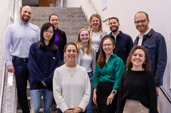 A diverse group of ten individuals stands on a staircase, smiling and posing together. They represent a collaborative research environment, likely related to an academic or scientific program, emphasizing teamwork and community within the postdoctoral network at DKFZ.
