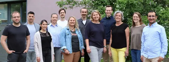 A diverse group of twelve people stands together outdoors, smiling. They are arranged in two rows, with greenery and a building in the background. The individuals appear to be colleagues, likely from a research or academic setting focused on cancer metabolism and microenvironment.