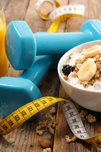 905879308 Eine Schüssel mit Müsli, Bananenscheiben und Beeren, daneben eine grüne Apfel, eine Orange, Hanteln und ein Maßband auf einem Holztisch.