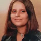 A young girl with shoulder-length brown hair smiles softly at the camera. She wears a black leather jacket and a simple necklace, set against a neutral background. The image conveys a warm and approachable demeanor.