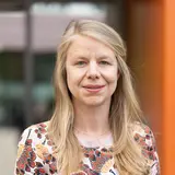 Porträt einer Frau mit langen, blonden Haaren, die ein bunt gemustertes Oberteil trägt. Sie steht vor einem unscharfen Hintergrund, der orangefarbene und graue Elemente zeigt. Die Frau lächelt freundlich und blickt direkt in die Kamera.