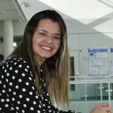 A smiling young woman with long hair, wearing a black and white polka dot blouse, leans on a railing in a brightly lit indoor setting. She wears glasses, conveying a friendly and approachable demeanor. The background shows a glimpse of a modern interior space.